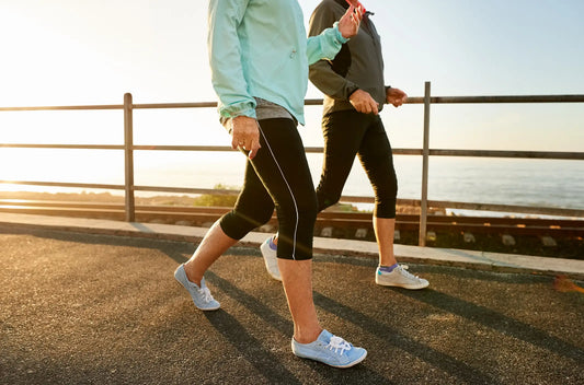 two women, visible from the shoulders down, wearing windbreakers and leggings and walking sneakers, walking along a boardwalk by the sea